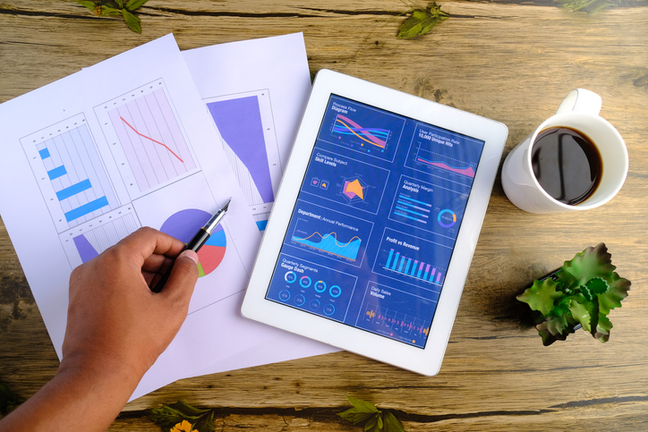 Electronic tablet with graphs, charts, metrics, etc. on top of papers with metrics on wooden background next to plant and coffee